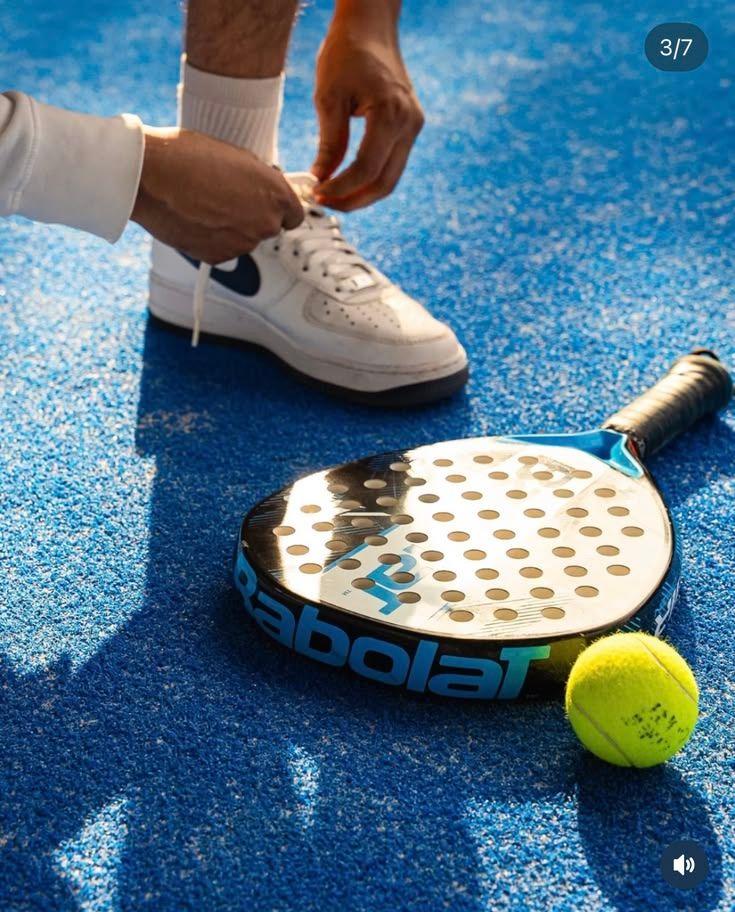 Joueur laçant sa chaussure sur une piste de padel, raquette et balle posées au sol